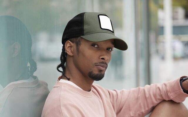 A man sitting up against a wall wearing a patch trucker cap